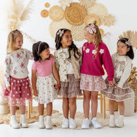 Five young girls in stylish outfits standing together in a decorative setting with floral and woven elements.