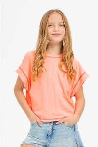 A young girl with long blonde hair wearing a coral orange short-sleeve t-shirt and light blue denim shorts, standing against a plain white background.