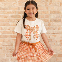 A young girl smiling in front of a light brick wall, wearing a cream short-sleeve t-shirt with a large orange and white gingham bow graphic on the front, paired with a matching orange and white gingham tiered skirt with a brown belt.