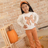 A young girl stands in a fall-themed room wearing a cream sweatshirt with a large orange and white plaid bow design on the front, paired with orange pants and a matching headband. She is posed next to a wooden crate holding a pumpkin against a brick wall background.