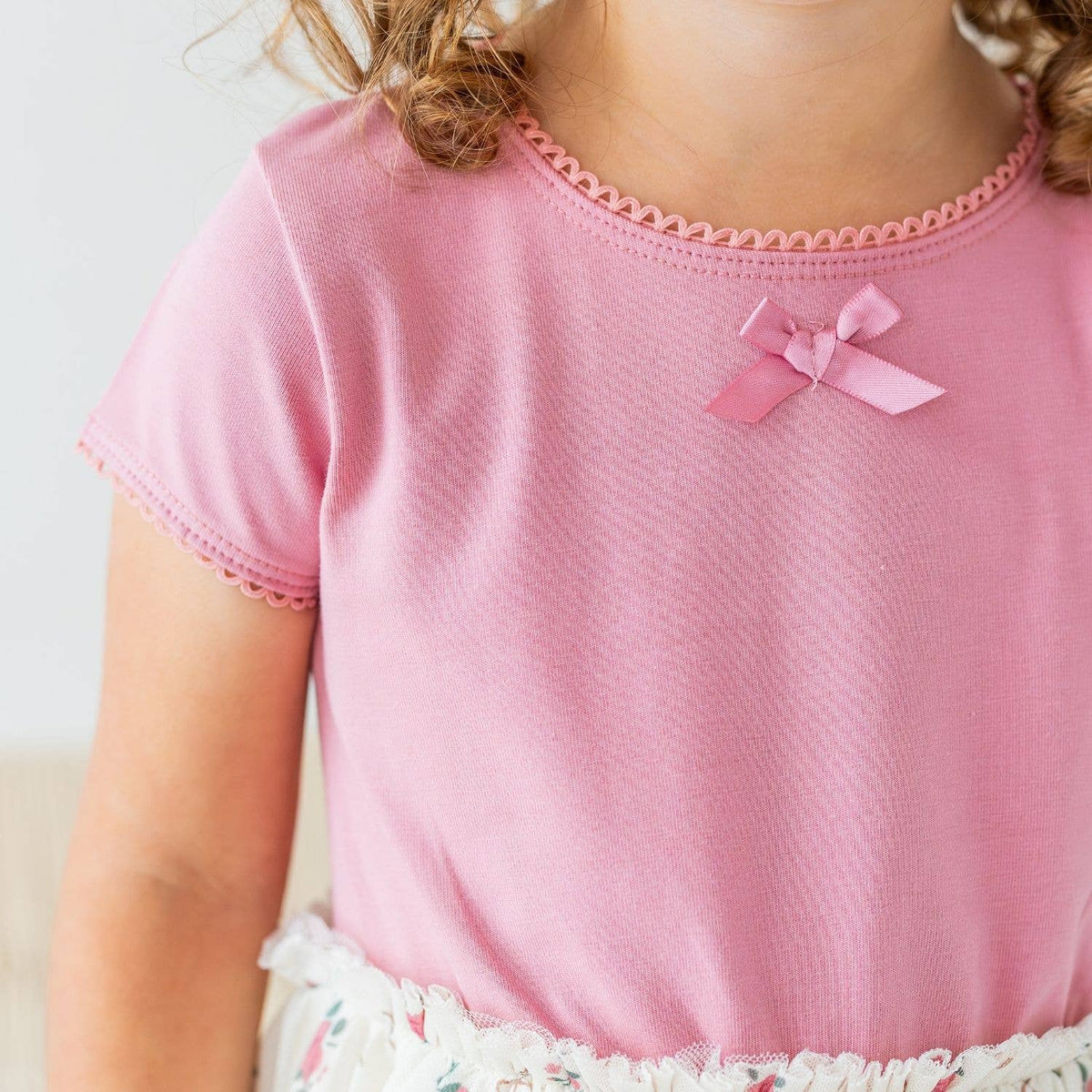 A close-up of a child wearing a light pink short-sleeve top with delicate pink scalloped trim around the neckline and sleeve cuffs, plus a small pink bow at the center chest.