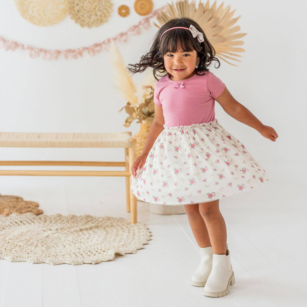 A young girl wearing a pink short-sleeve top and a white flared skirt with pink floral prints, posing in a bright, decorated room with a woven bench and pampas grass.