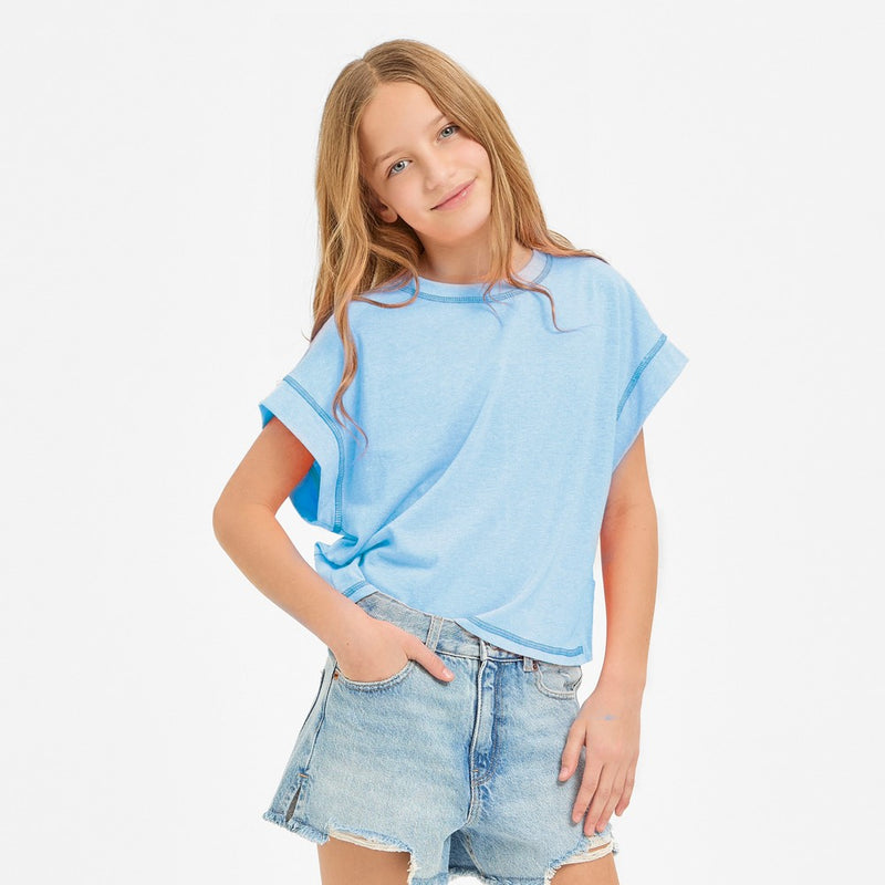 A girl wearing a neon coral sleeveless boxy crop top with a crew neckline and visible contrast overlock stitching along the armholes, side seams, and hem, paired with light-wash denim shorts.