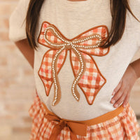 A close-up of a child wearing a cream-colored t-shirt with a large orange and white gingham bow applique outlined with faux pearls and brown trim. The child is also wearing a matching orange and white gingham skirt with a brown ribbon tie.