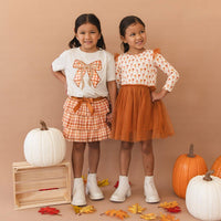 Two young girls modeling autumn-themed outfits in a studio setting with pumpkins and fall leaves. The girl on the left wears a white graphic T-shirt with an orange bow print, a gingham skirt, and white boots.