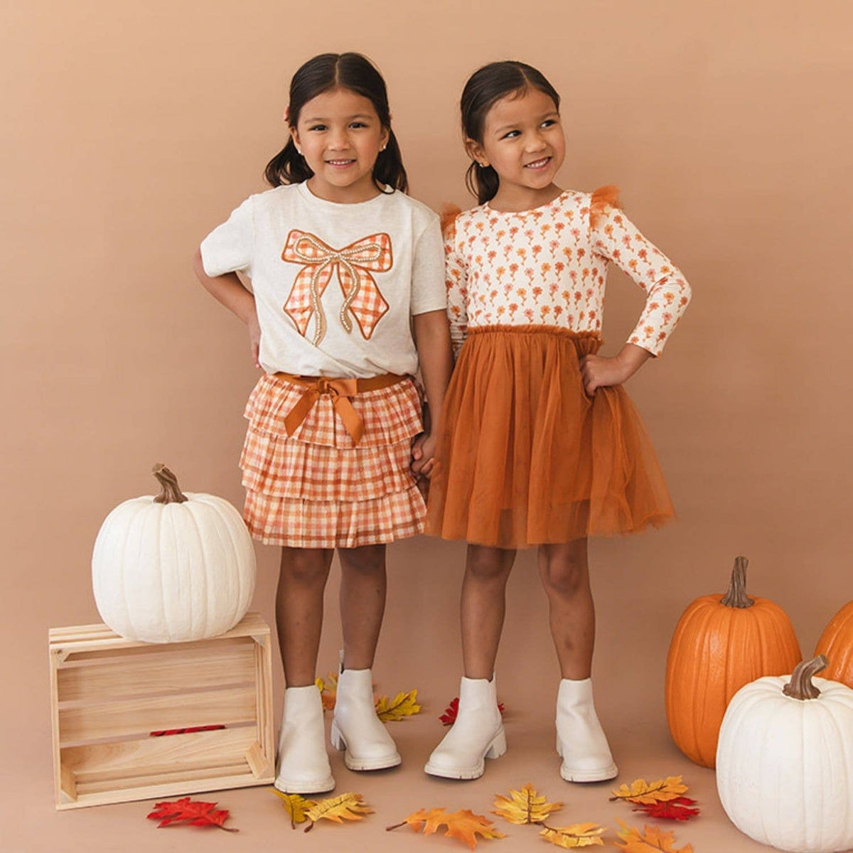 Two young girls modeling autumn-themed outfits in a studio setting with pumpkins and fall leaves. The girl on the left wears a white graphic T-shirt with an orange bow print, a gingham skirt, and white boots.