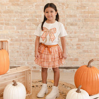 A young girl stands in a fall-themed studio setting wearing a white t-shirt with an orange and white gingham bow print, a matching orange and white gingham pleated skirt, and white ankle boots. She is posed between pumpkins in front of a light brick wall.
