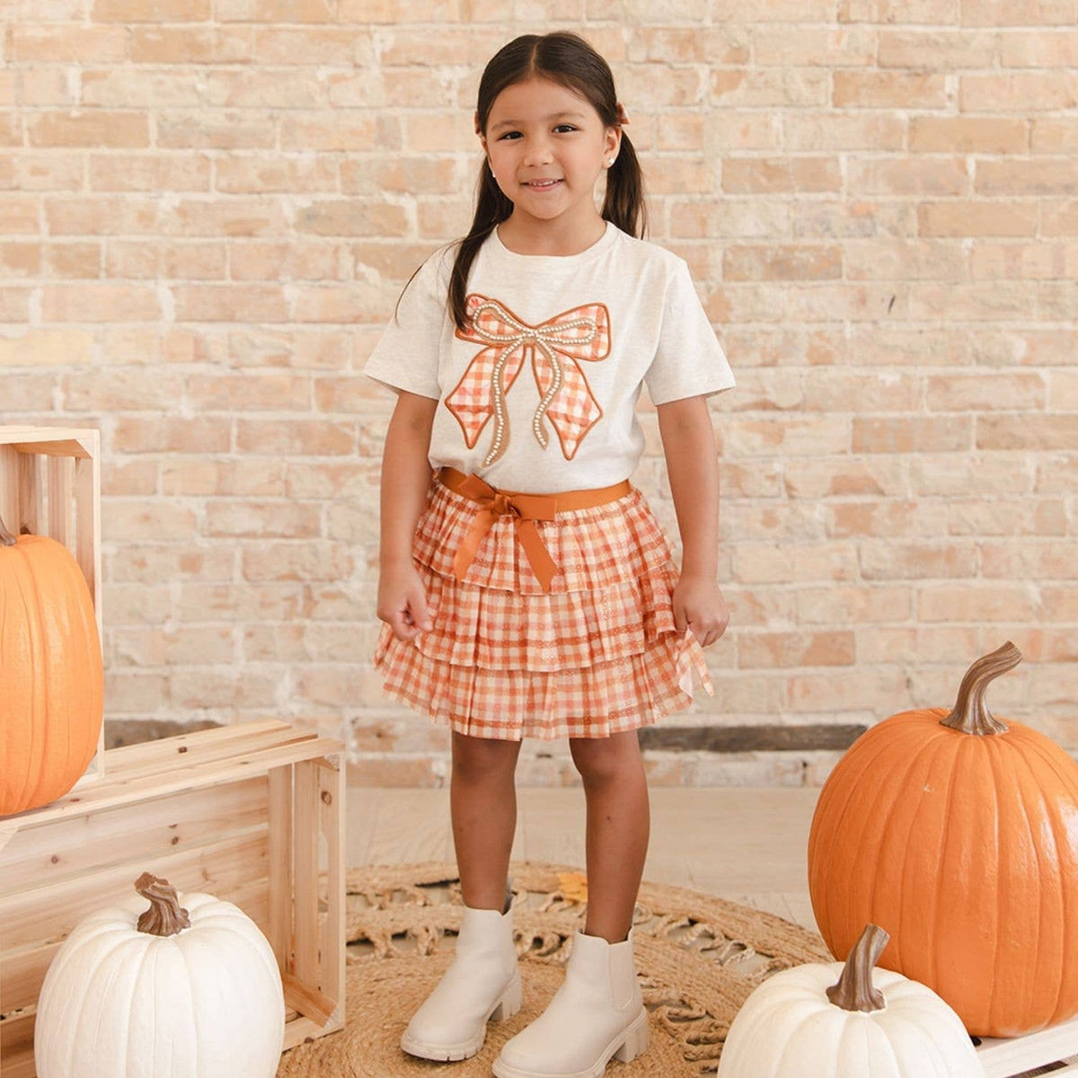A young girl stands in a fall-themed studio setting wearing a white t-shirt with an orange and white gingham bow print, a matching orange and white gingham pleated skirt, and white ankle boots. She is posed between pumpkins in front of a light brick wall.