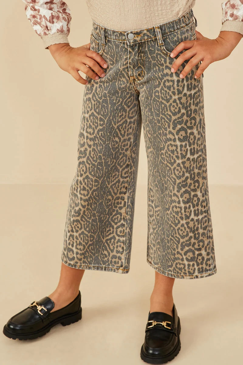 A person wearing cropped wide-leg denim jeans with an allover leopard print pattern in beige and gray, paired with black loafers and a cream top, shown against a plain beige studio background.