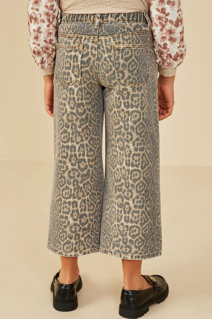 Back view of a person wearing wide-leg cropped denim jeans with an allover leopard print pattern in beige, gray, and brown tones, paired with a floral long-sleeve top and black loafers, against a plain beige studio background.