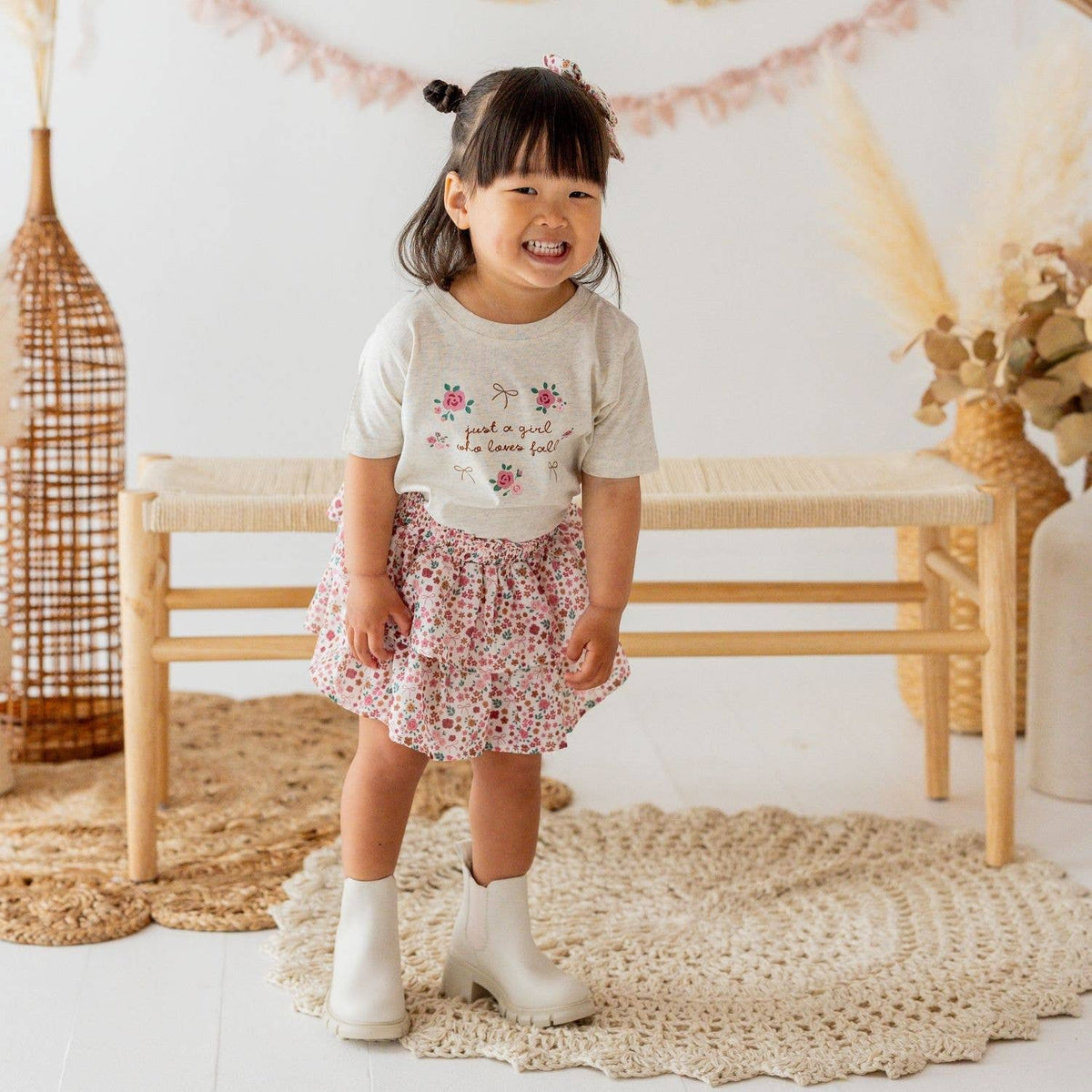 Child wearing a floral skirt and white boots standing on a textured rug with a neutral background.