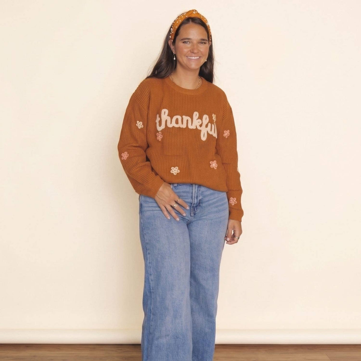 A woman wearing a rust-orange ribbed knit sweater with 'thankful' written across the chest in white script, paired with light-wash blue jeans and a matching orange headband, standing against a plain cream wall.