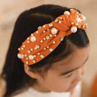 An orange fabric twist-front headband decorated with white faux pearls and gold rhinestone accents, shown worn by a young girl.