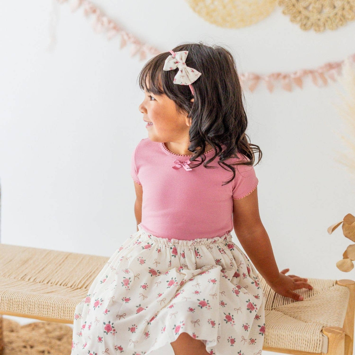 Child wearing a pink top and floral skirt with a bow in hair, standing indoors.