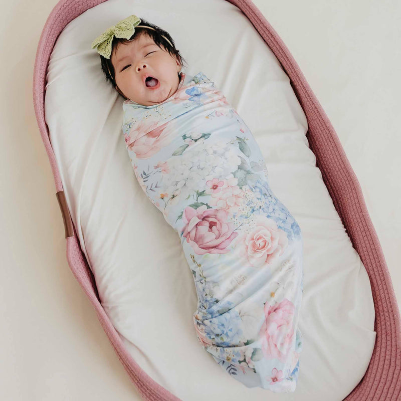 A newborn baby swaddled in a light blue floral swaddle blanket, lying in a pink bassinet. 