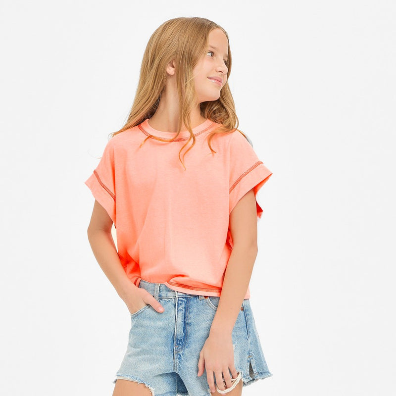 A girl wearing a neon coral sleeveless boxy crop top with a crew neckline and visible contrast overlock stitching along the armholes, side seams, and hem, paired with light-wash denim shorts.
