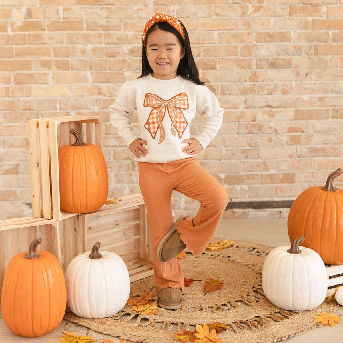 A young girl stands in a fall-themed studio setting wearing a cream sweatshirt with an orange gingham bow graphic, matching orange pants, and an orange gingham headband. The background features a brick wall, wooden crates, pumpkins, and a jute rug.