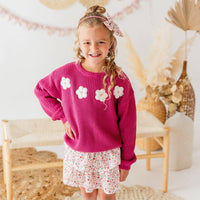 Young girl wearing a pink sweater with white floral patterns and a matching skirt in a decorative indoor setting.